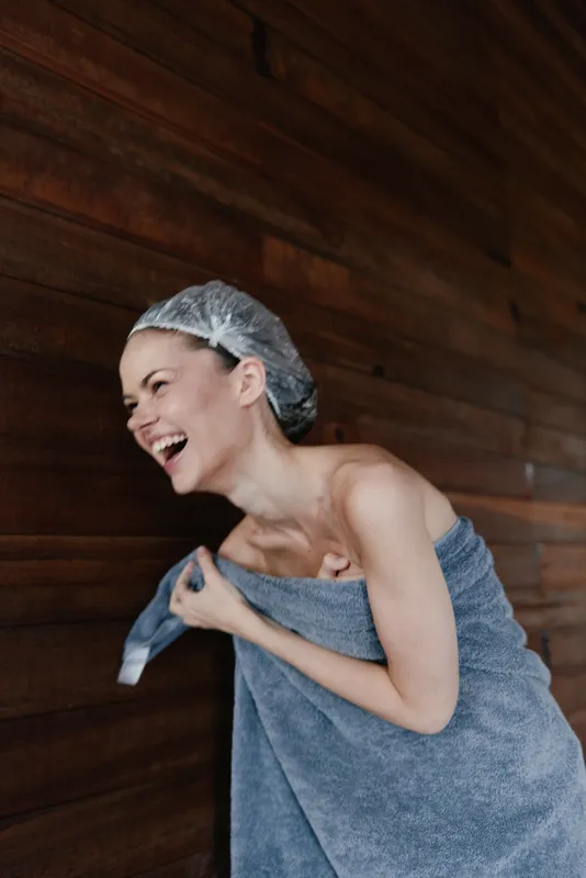 laughing woman wrapped in blue towel and shower cap standing near wooden wall, happiness and freshness after bath, natural skin care moment indoors