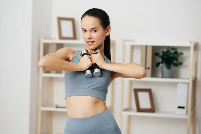 Fitness woman performing arm workout at home with dumbbells, wearing a grey sports bra and leggings, focused expression, modern living room backdrop with shelves and a plant.