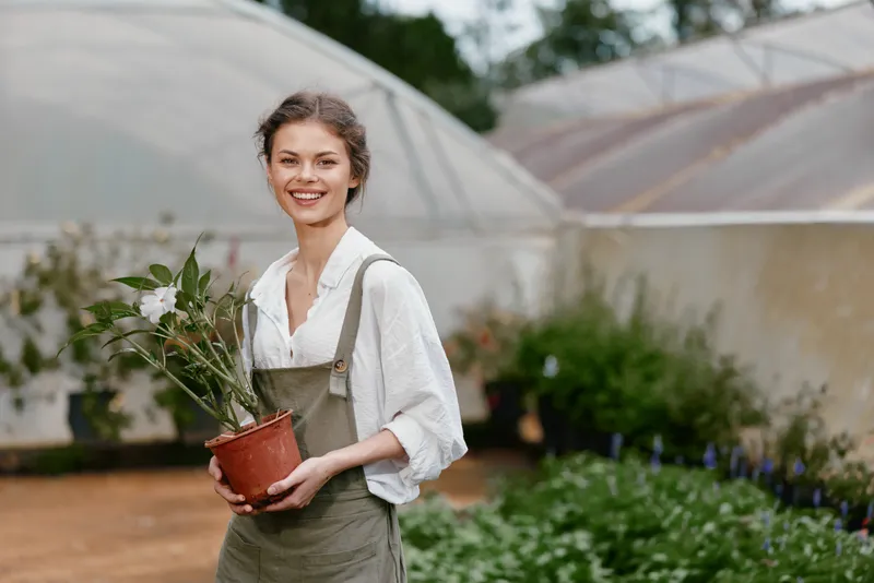 Young woman smiling in a greenhouse, holding a potted plant, wearing a light shirt and overalls, surrounded by greenery and vibrant flowers