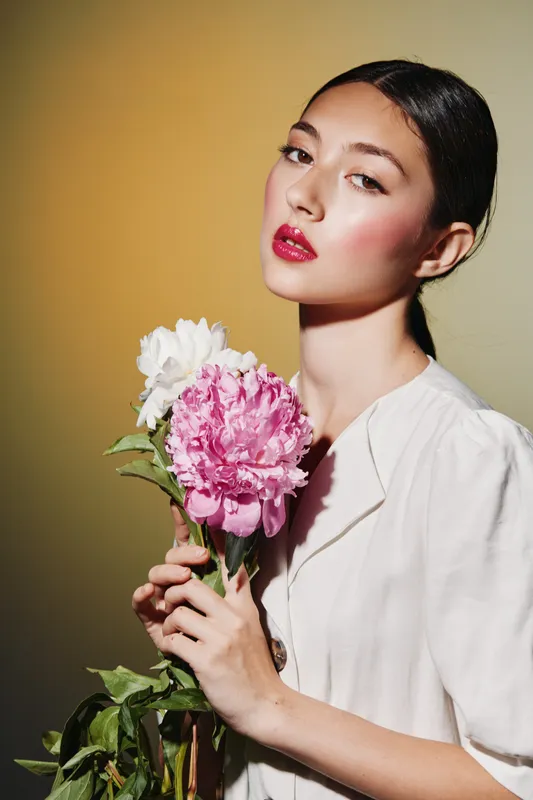 Young woman with flawless skin and pink makeup poses holding peonies against a soft pale background. Fashion creative beauty concept with natural gentle lighting and elegance.