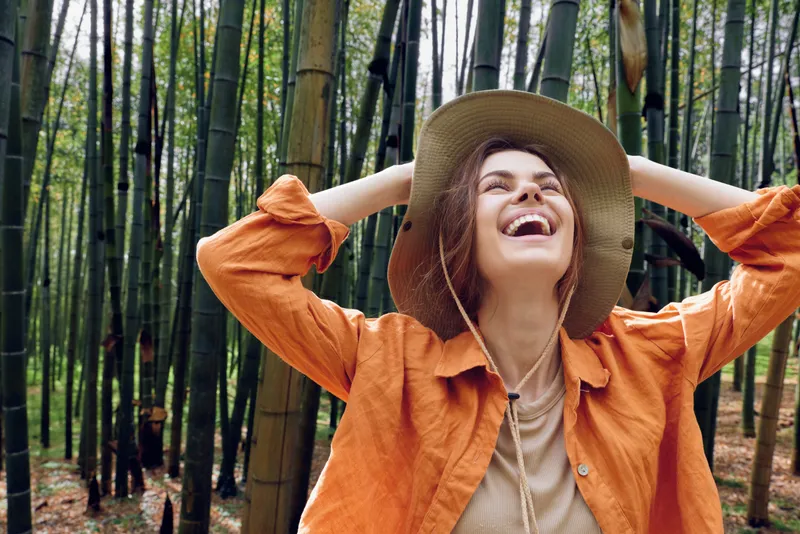 Woman in hat laughing in a bamboo forest, nature portrait filled with joy and happiness; casual traveler smiling and carefree among green trees and sunlight.