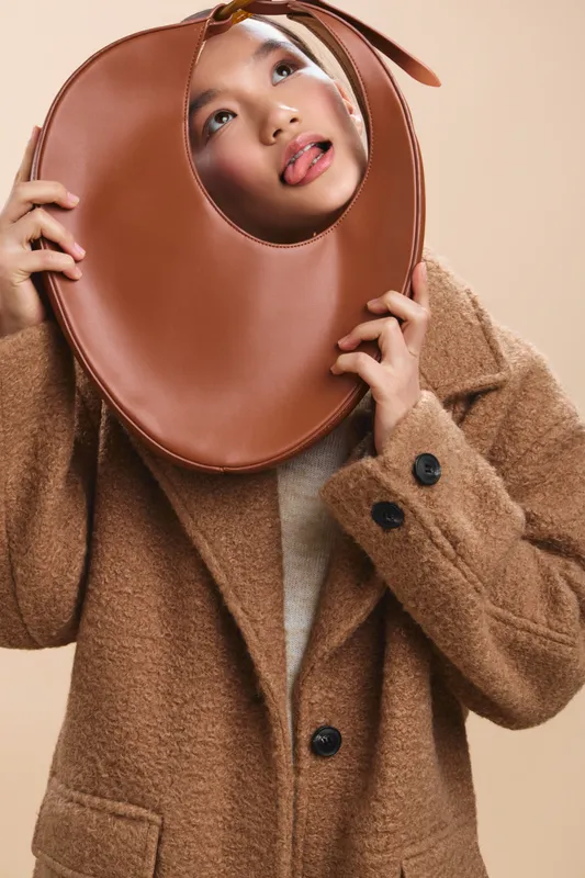 Asian woman wearing a warm textured coat holds a large circular accessory against her face, creating a playful fashion portrait with soft lighting and a neutral studio backdrop.