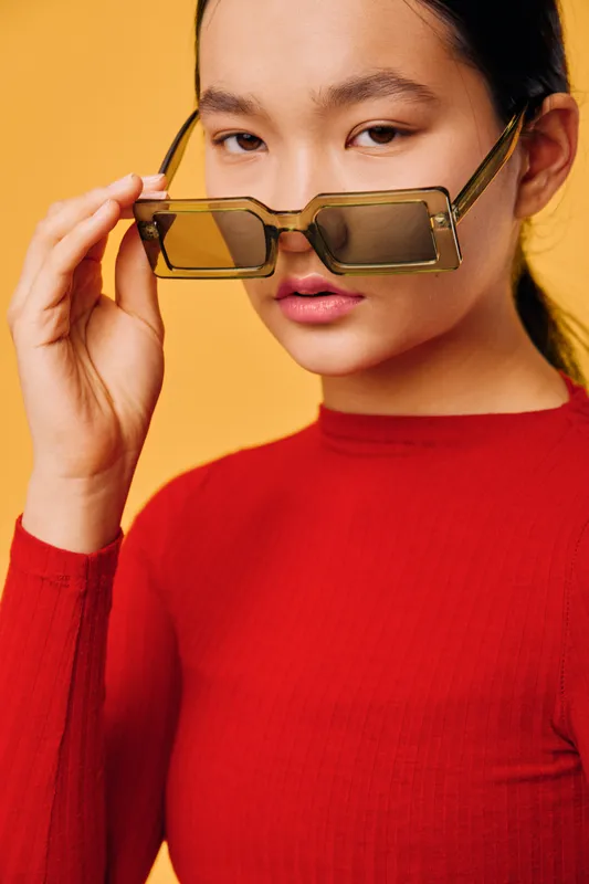 Woman portrait adjusting sunglasses with hand, wearing red top against yellow background. Closeup fashion model showcasing stylish eyewear and confident expression indoors.