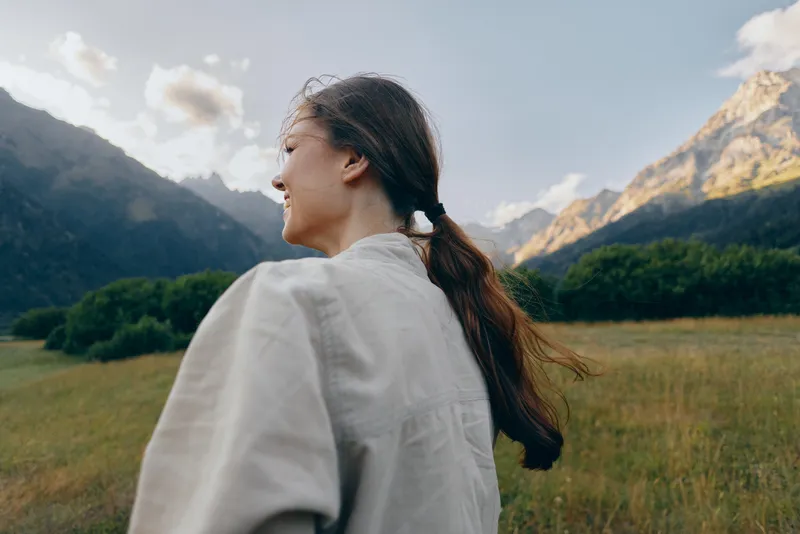 Smiling woman with long hair outdoors in nature, wearing casual clothes, enjoying fresh air, mountain landscape background, peaceful and relaxed mood