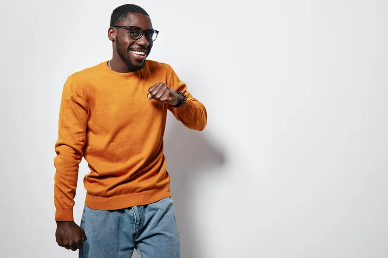 African American man with dark skin wearing orange sweater and glasses smiling and dancing against white background in casual style and joyful mood.