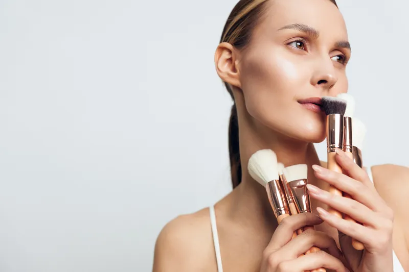 Young woman holding makeup brushes near face with natural skin and subtle makeup. Concept of beauty routine, cosmetics, and skincare with soft lighting on white background.
