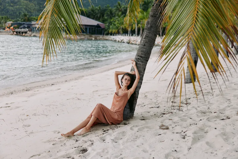 Relaxed woman in a summer dress sitting on the sandy beach near a palm tree with tropical background and calm sea water.
