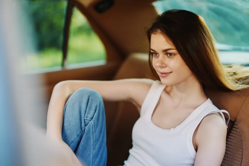 Young woman in casual attire relaxing in a car, smiling and enjoying a sunny day, with soft natural light highlighting her features and relaxed expression.