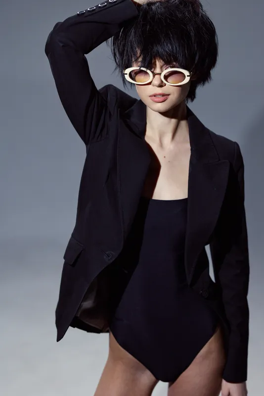 Stylish woman with black jacket and swimsuit wearing white sunglasses in studio lighting.