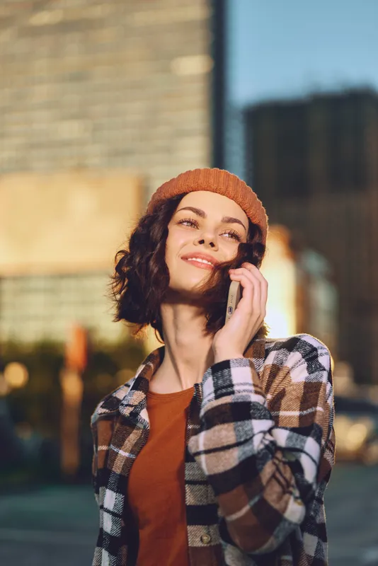 woman phone urban golden smile candid lifestyle beanie: Young woman in a beanie talks on mobile on a city street at golden hour glow, authentic mindful living and emotional storytelling atmosphere.