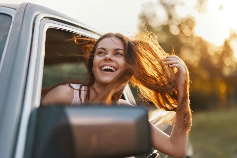 Young woman enjoying carefree moments with flowing hair in the open window of a car, capturing joy and freedom in a warm sunset background.