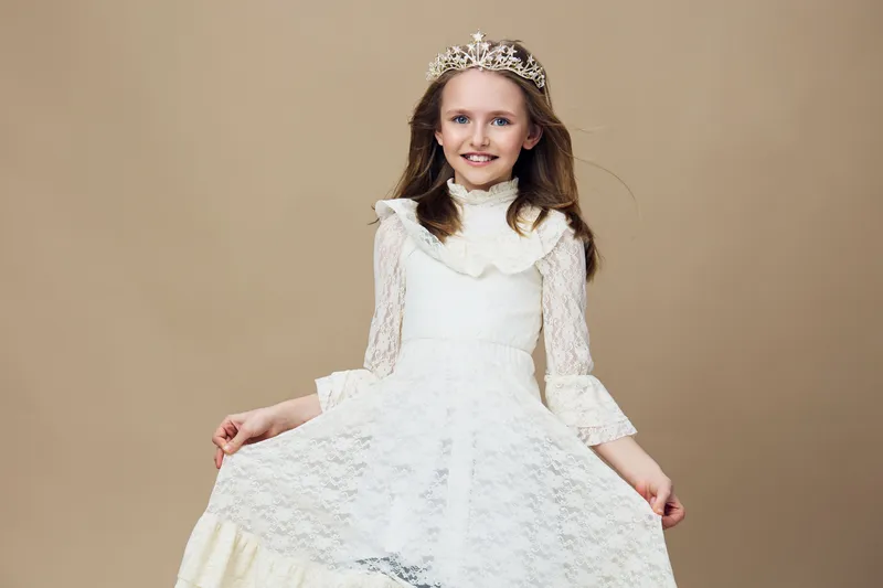 Young girl in an elegant white dress and a tiara, smiling beautifully against a neutral beige background, perfect for portraits, celebrations, and childhood themes