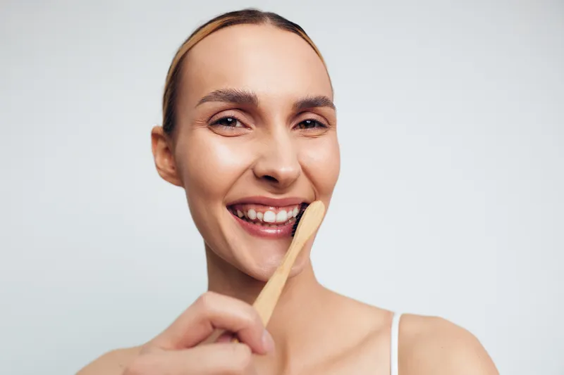 A young woman with brunette hair smiles while brushing her teeth with a bamboo toothbrush against a plain white background, showing a natural and eco-friendly morning routine.