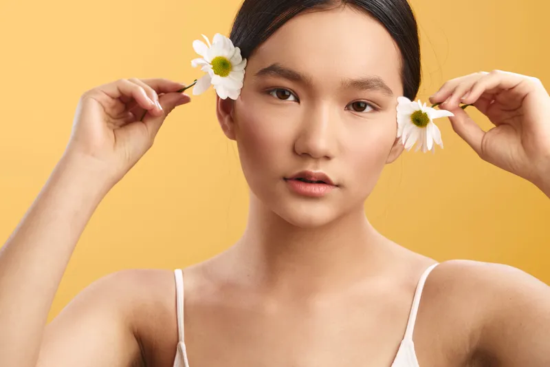 Asian woman with a delicate expression holds flowers near her ears, creating a serene portrait in a warm studio setting that emphasizes beauty, balance, and graceful poise, conveying calm confidence