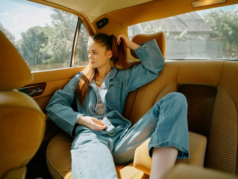 A young woman in a denim outfit sitting in a vintage car, exuding a relaxed vibe with a serene expression, captured in bright, natural light.