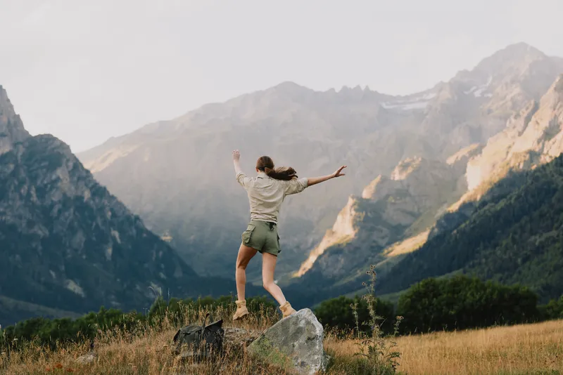 A woman stands on a rock in an open field, arms raised, with mountains and sky behind. Outdoor life and freedom are the themes, hinting at adventure and resilience.