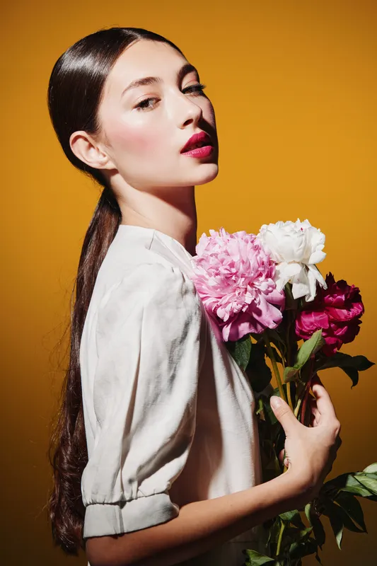 Fashion model with long dark hair poses with pink and white flowers against orange background. Soft studio lighting accentuates makeup, skincare, and creative beauty concepts.