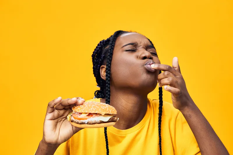 Hungry woman enjoying a delicious burger against a vibrant yellow background, showcasing joy and satisfaction in her expression, perfect for food related content