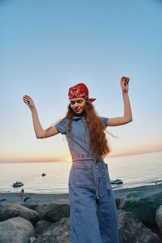 Lifestyle woman in modern boho-western denim street style with long hair and red bandana enjoys sunset by the sea in warm film color tones.
