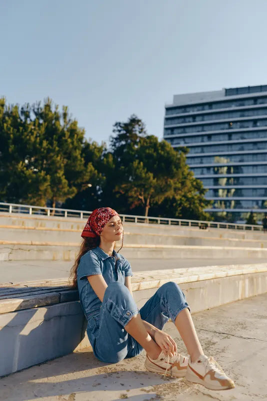 Lifestyle woman in modern boho-western denim street style sitting outdoors, relaxed pose, film color, casual summer fashion, urban background, sunny day.