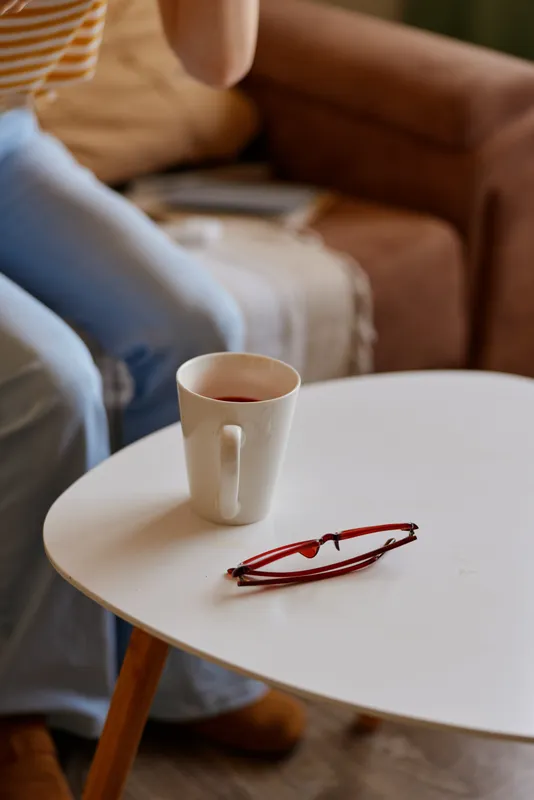 Cozy living room scene with a white mug on a small round table and glasses nearby, a blurred figure on a sofa suggesting a woman enjoying a quiet morning.
