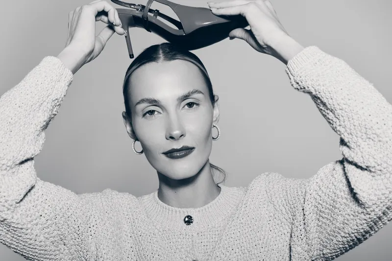Caucasian woman with fair skin and red lips holds a high heel shoe above her head. Studio portrait in black and white with confident facial expression.
