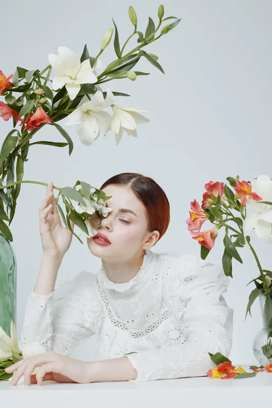 Elegant model with a serene expression, draped in a white vintage dress, surrounded by vibrant flowers, creating a soothing and artistic atmosphere