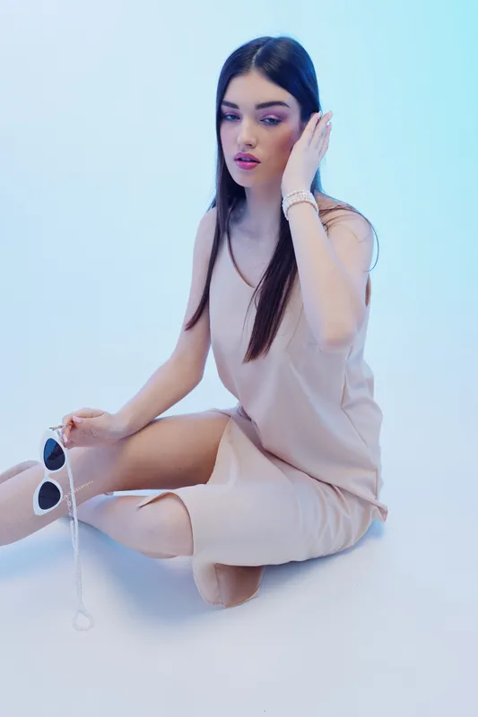 Young woman in beige dress sits on the floor holding sunglasses. She has long dark hair and subtle makeup. Studio shot with soft lighting. Concept of woman beauty fashion.