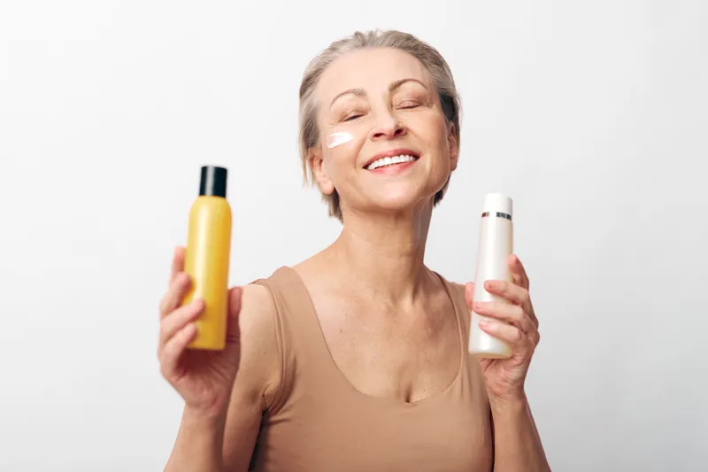 Happy mature woman with short hair holding two cosmetic bottles and applying face cream on cheek. Skincare concept with natural beauty, self-care, wellness, and anti-aging on white background.