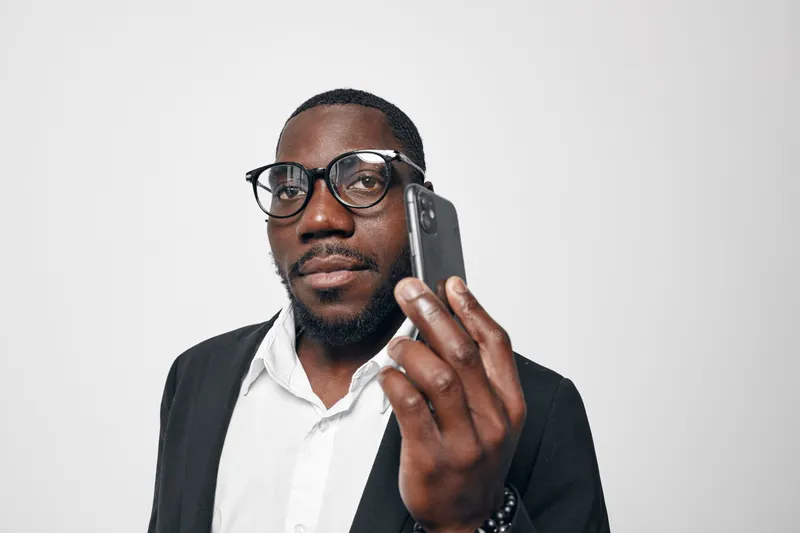 African American man with dark skin wearing glasses and white shirt holding smartphone close to face for communication or call on white background. Technology, portrait and business concept.