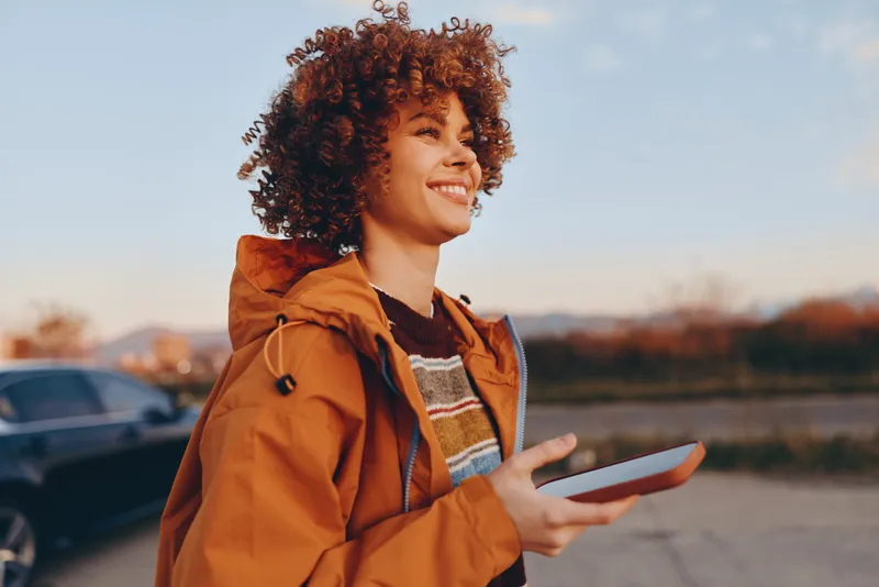 woman lifestyle outdoors portrait smiling curly hair in orange jacket holding smartphone near parking road at sunset, travel communication, cheerful mood, natural light, soft focus