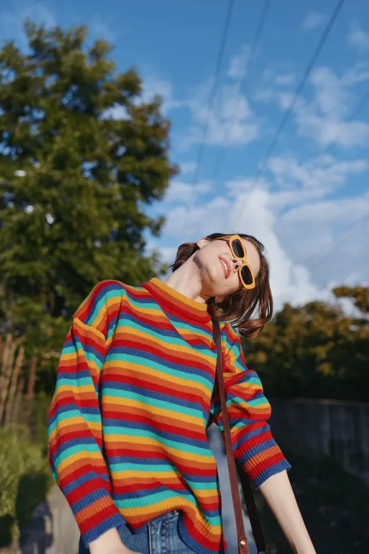 Woman in a colorful striped sweater wearing yellow sunglasses, smiling and leaning back outdoors in sunlight. Casual outfit with denim jeans and braided hair, relaxed suburban lifestyle portrait.