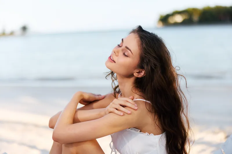 Portrait of a young woman sitting on a sunny beach with eyes closed and wind in her hair, wearing a light sundress, radiating calm and natural beauty by the sea.