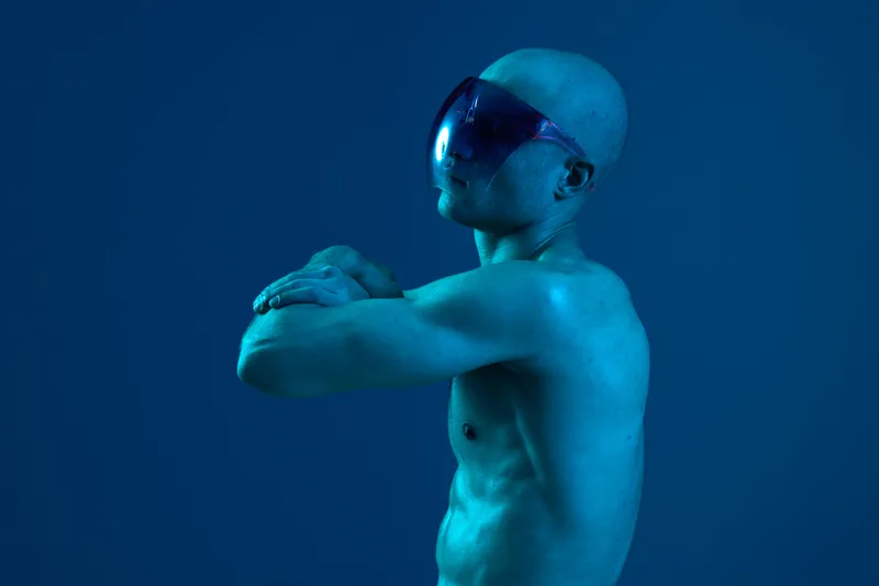A man showcasing a strong pose in a unique setting, exuding confidence and charisma. The blue lighting emphasizes his features, creating a striking black and white image.