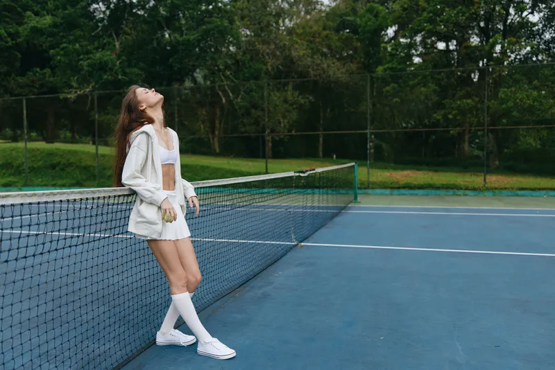 Young female tennis player in sporty attire exudes confidence on the tennis court, surrounded by lush greenery, showcasing determination and a healthy lifestyle