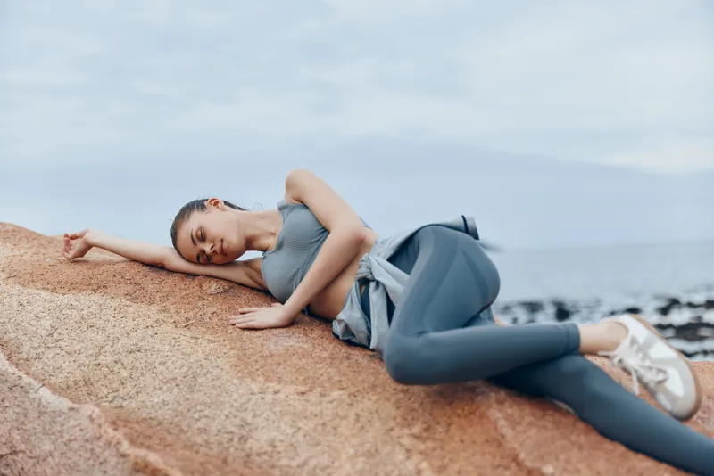Young woman in activewear relaxing on rocky beach, showcasing a serene expression and healthy living concept with muted colors