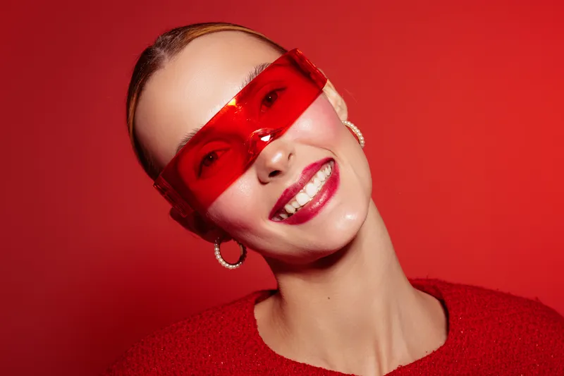 Smiling woman with red sunglasses and red sweater poses against red background. Bright makeup and hoop earrings create a bold, modern fashion portrait with vibrant color contrast.