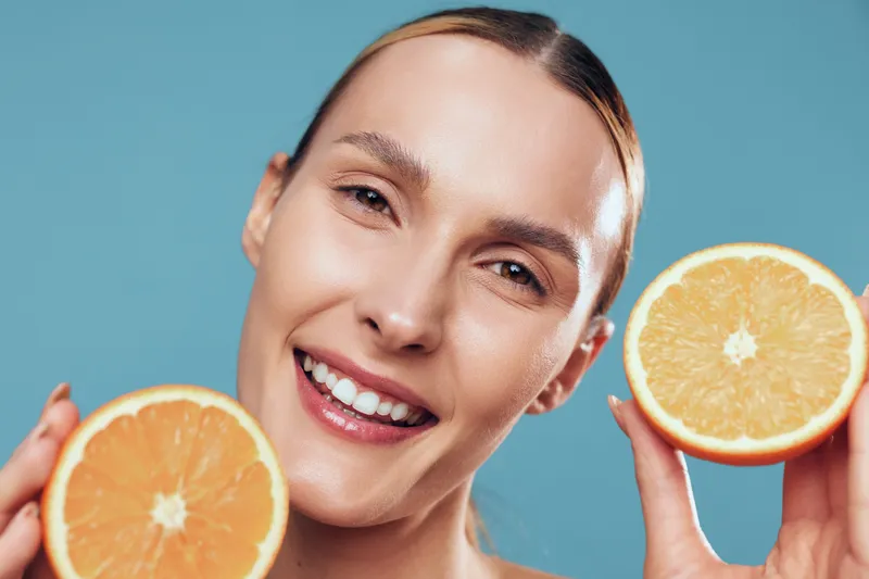 Young woman with slicked-back hair smiles joyfully while holding two halves of a fresh orange near her face against a blue background.