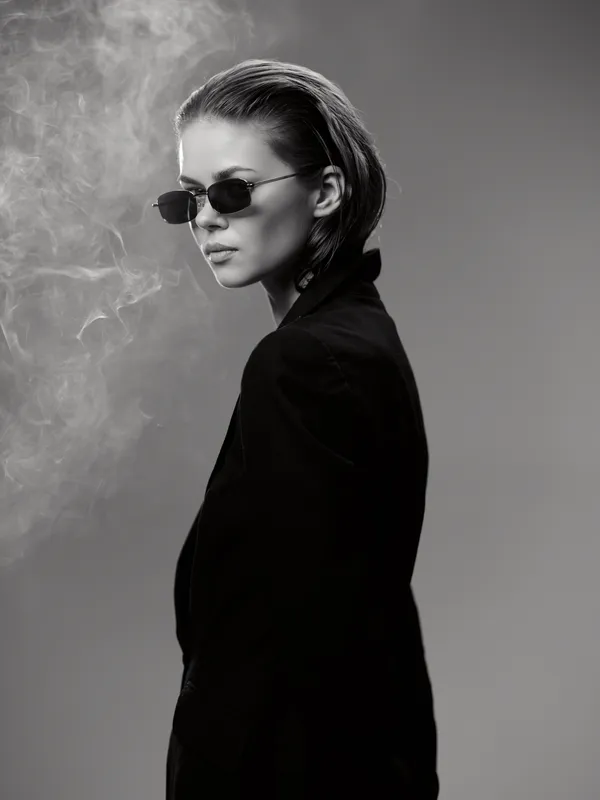 Black and white portrait of a stylish woman wearing sunglasses and a dark jacket with smoke in the background.