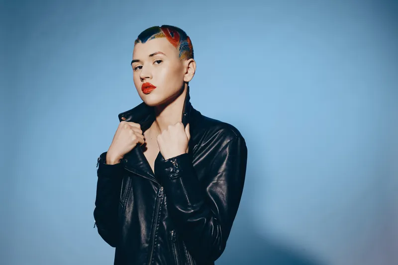 woman with colorful hair and leather jacket posing confidently against blue background, bold makeup, edgy style, artistic urban fashion portrait.