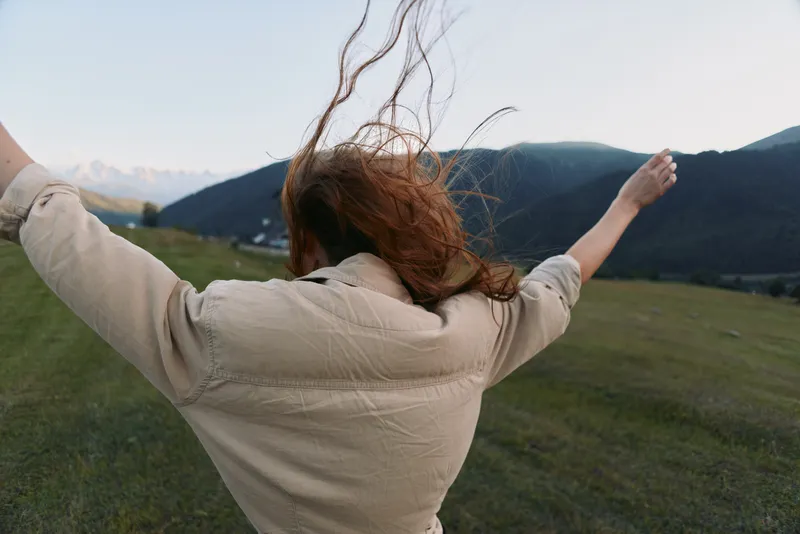 Woman in beige jacket raises arms in a windy countryside, embracing nature and freedom, a wide meadow and distant mountains under a bright sky, a pure joyful outdoor moment
