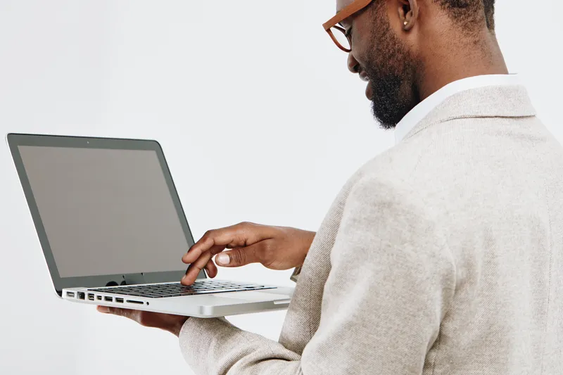 Professional man using laptop computer, side view, wearing beige sweater, with beard and glasses, working indoors with plain white background. Technology work and remote office concept.
