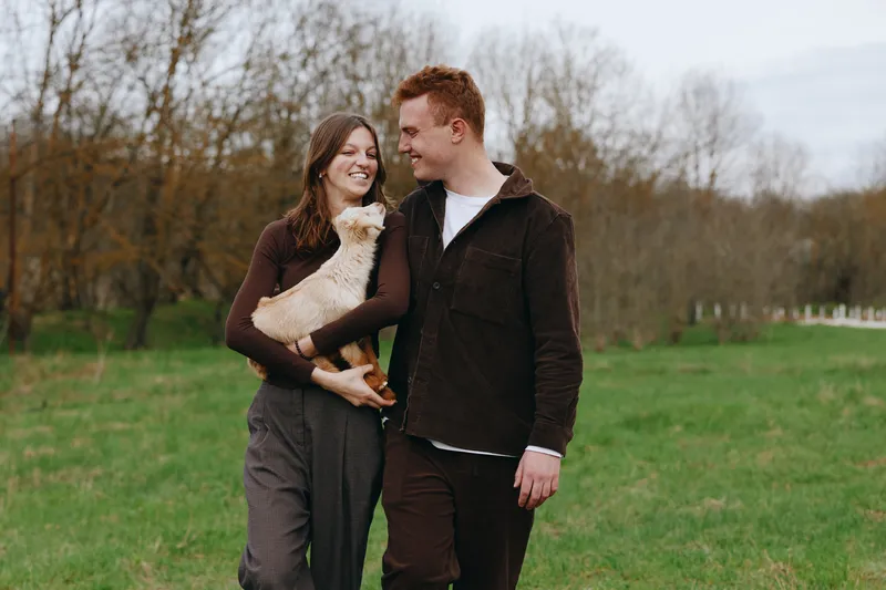 Playful couple walking in a green park with a small dog, smiling and enjoying time together, casual autumn attire, candid lifestyle moment, serene outdoor scene.
