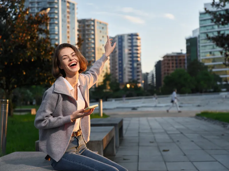A cheerful professional woman sits on a concrete bench in an urban park, smiling broadly and waving with one hand while holding a smartphone in the other, enjoying a bright moment outdoors.