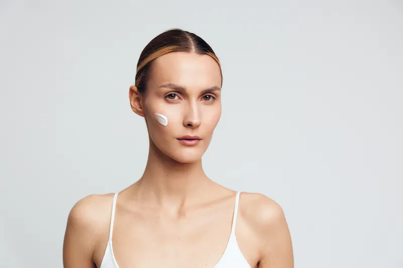 Young woman with clear skin applying moisturizer cream on face, neutral background, natural beauty, skincare routine, focused expression, simple white top, minimal makeup.