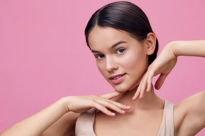 Young woman with a serene expression in a light beige top, showcasing a natural beauty look against a soft pink background, highlighting self confidence and femininity