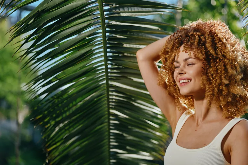 Smiling woman with curly hair enjoys sunlight outdoors near tropical palm leaves, wearing a white tank top in a natural green environment on a sunny day.