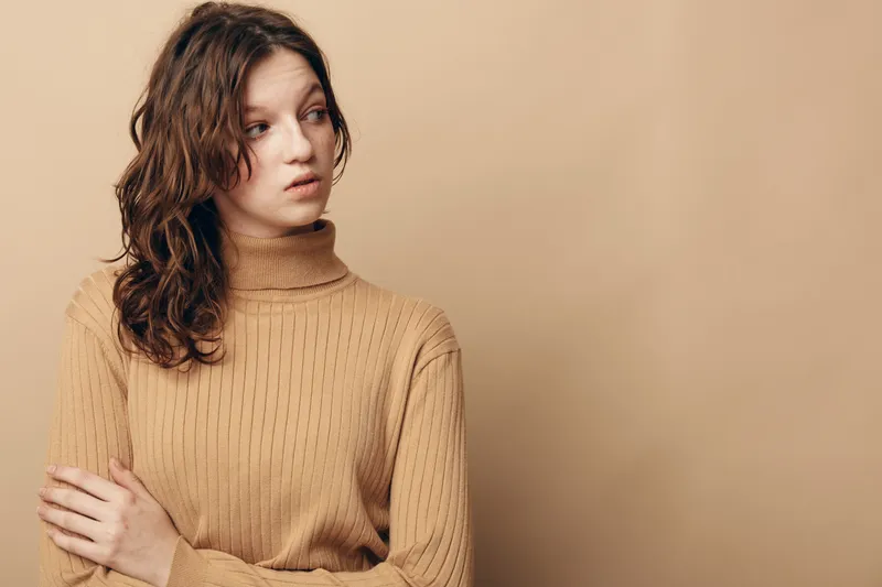 Young woman with curly hair in beige sweater looks away thoughtfully on beige background. Concept of contemplation, emotion, and casual fashion portrait.