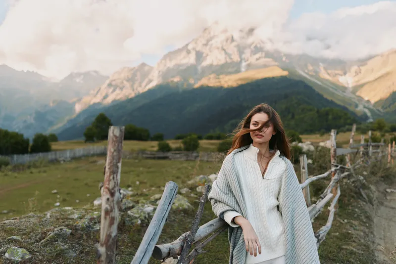 A young woman in a cozy knit poncho stands beside a wooden fence in a mountain valley, gazing ahead with a calm, reflective mood.