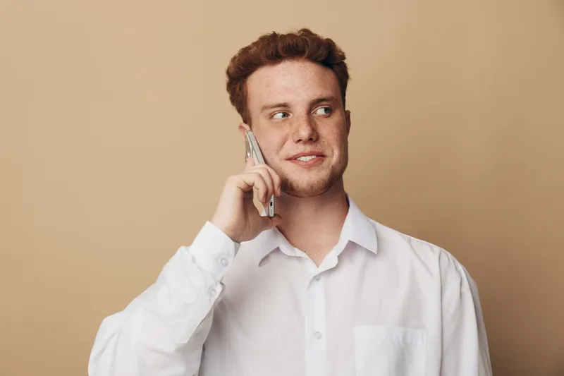 Young man with red hair wearing white shirt talks on smartphone looking away with slight smile. Neutral beige background. Concept of communication, technology, and casual lifestyle.